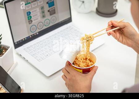 Eine Person isst Instant-Nudeln an ihrem Schreibtisch und hebt die Nudeln mit Stäbchen aus einer Tasse. Stockfoto