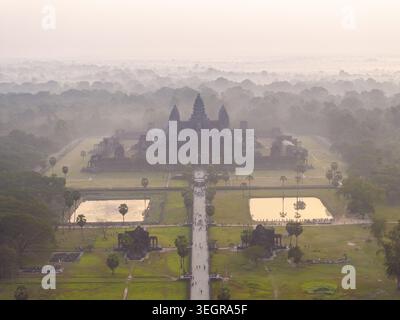 Blick aus der Vogelperspektive auf den alten Tempelkomplex Angkor Wat, der in Morgennebel gehüllt ist, mit seinen berühmten Steintürmen und dem umliegenden üppigen Wald in Krong Siem R Stockfoto