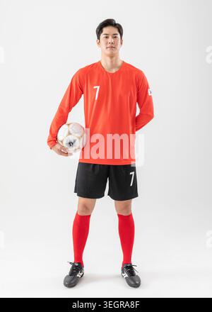 Ein junger asiatischer Fußballspieler steht selbstbewusst in einem weißen Studio, der einen Fußball hält. Stockfoto