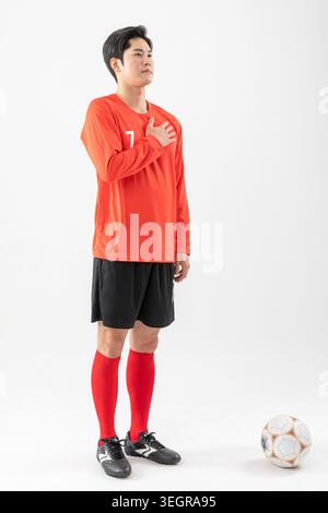 Ein junger asiatischer Fußballspieler steht mit der Hand auf dem Herzen und bereitet sich darauf vor, die Nationalhymne zu singen. Stockfoto