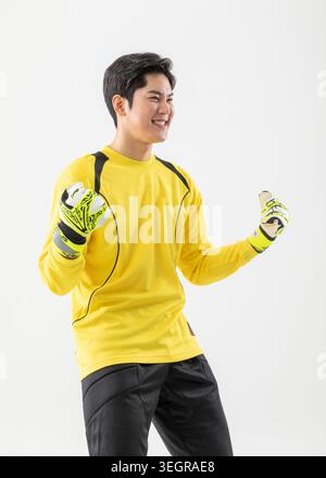 Ein junger asiatischer Torhüter steht mit triumphalen Armen in einem gelben Trikot und passenden Handschuhen vor weißem Hintergrund. Stockfoto