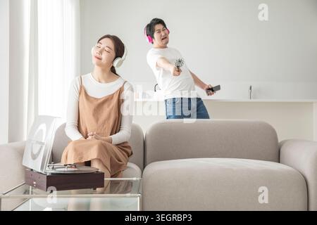 Eine junge Frau sitzt auf einer Couch und hört Musik auf einem Plattenspieler, während ihr Freund im Hintergrund tanzt. Stockfoto
