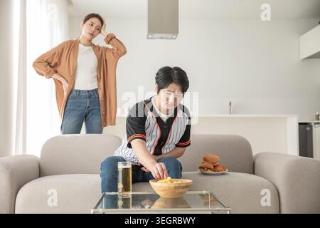 Ein junger Mann sitzt auf einer Couch, isst Chips und schaut fern, während seine Freundin im Hintergrund steht und uninteressiert aussieht. Stockfoto