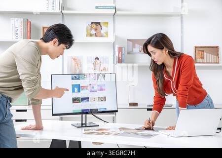 Ein Mann und eine Frau arbeiten zusammen an einem Schreibtisch, beide konzentrieren sich auf einen Computerbildschirm, auf dem eine Bilderbibliothek angezeigt wird. Stockfoto