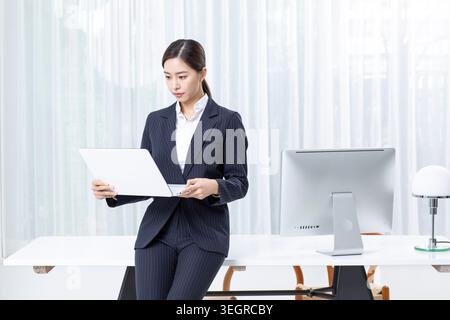Eine Frau in einem Business-Anzug steht in einem Büro, hält einen Laptop und sieht sich ein Dokument an. Stockfoto