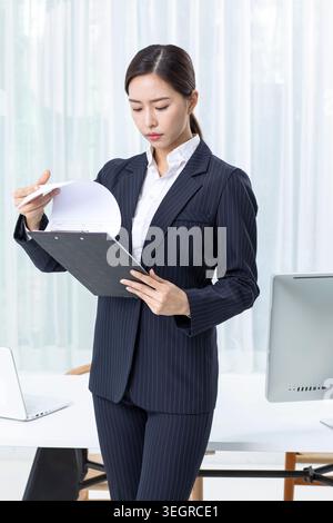 Eine junge asiatische Frau im Business-Anzug sitzt an einem Schreibtisch und studiert intensiv ein Dokument. Stockfoto