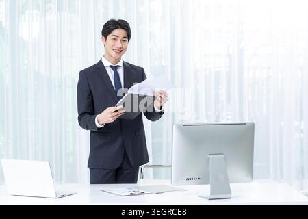 Ein junger asiatischer Geschäftsmann im Anzug lächelt und hält in einem modernen, gut beleuchteten Büro eine Aktenmappe. Stockfoto