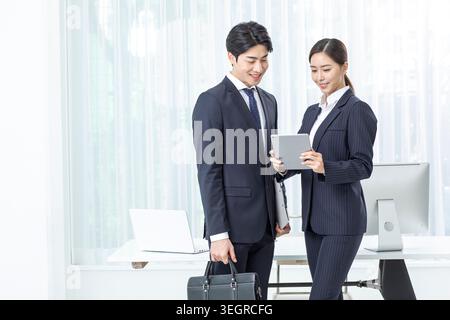 Eine junge asiatische Geschäftsfrau und ein junger asiatischer Geschäftsmann in einem modernen Büro, beide lächeln, während sie gemeinsam ein Tablet überprüfen. Stockfoto