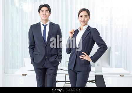 Zwei junge asiatische Geschäftsleute, ein Mann und eine Frau, stehen selbstbewusst in einem modernen Büro und lächeln in die Kamera. Stockfoto