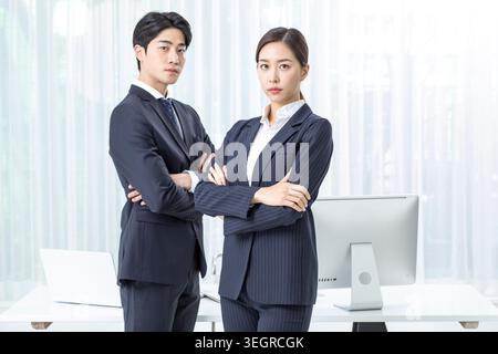 Zwei junge Profis in einem modernen Büro, die Rücken an Rücken mit überkreuzten Armen stehen und selbstbewusst und konzentriert aussehen. Stockfoto