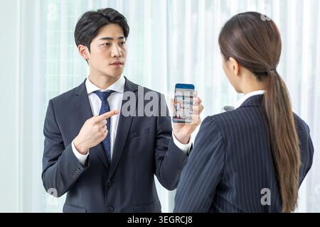 Ein junger asiatischer Mann in einem Anzug zeigt auf einen Börsendiagramm auf seinem Smartphone, während eine Frau in einem Business-Anzug es ansieht. Stockfoto
