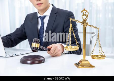 Ein junger Berufstätiger, der an einem Schreibtisch sitzt, einen Hammer hält, umgeben von Gesetzbüchern und einem Laptop, in einem modernen Anwaltsbüro. Stockfoto