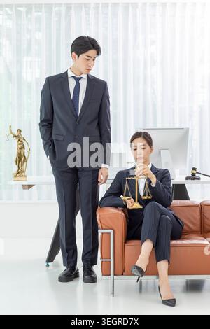 Ein junger asiatischer Mann und eine junge asiatische Frau in Geschäftskleidung stehen in einem modernen Büro, umgeben von Gesetzbüchern und juristischen Symbolen. Stockfoto