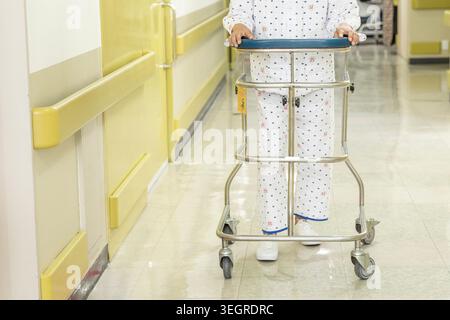 Ein Patient, der einen Krankenhausmantel trägt, benutzt einen Geher in einem Krankenhausflur. Stockfoto