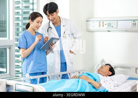 Zwei Ärzte und eine Krankenschwester in einem Krankenhauszimmer besprechen die Akte eines Patienten, während eine Krankenschwester die Vitalparameter des Patienten überprüft. Stockfoto