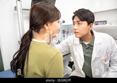 Eine junge Frau, die einen grünen Pullover trägt, sitzt in einer Arztpraxis und sieht einen Arzt an, einen männlichen Asiaten, in einem weißen Labormantel, während er sie untersucht. Stockfoto