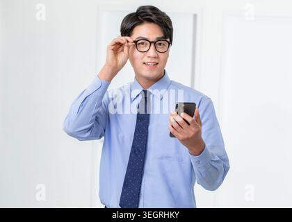 Ein junger Geschäftsmann im Anzug lächelt und passt seine Brille an, während er sein Handy in einem modernen Büro hält. Stockfoto