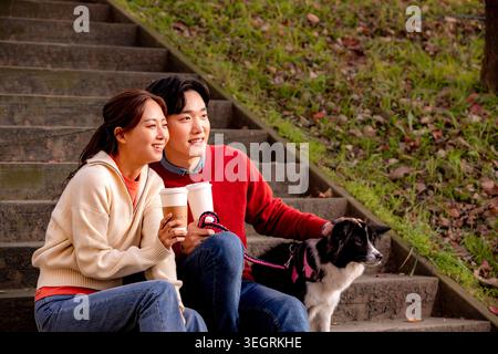 Ein junges Paar und ihr Hund sitzen auf einer Parkbank, lachen und genießen gemeinsam Kaffee. Stockfoto