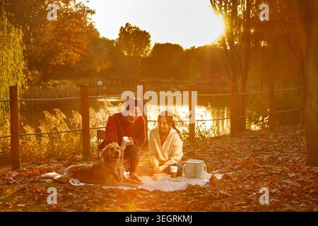 Ein junges Paar genießt an einem sonnigen Herbsttag ein Picknick am See mit seinem goldenen Retrieehund. Stockfoto