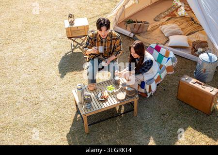 Ein junges Paar genießt ein Picknick auf einem sonnigen Campingplatz, umgeben von einem Zelt, Campingausrüstung und einem Tisch mit Speisen und Getränken. Stockfoto