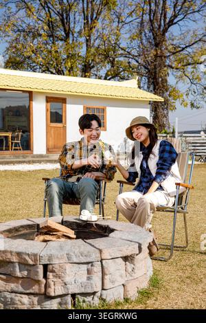 Ein Mann und eine Frau sitzen zusammen am Lagerfeuer und trinken mit einer Flasche Wein in einer sonnigen, ländlichen Landschaft. Stockfoto