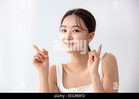 Eine junge asiatische Frau in einem weißen Tank zeigt mit beiden Zeigefingern nach links und blickt mit einem neutralen Ausdruck direkt in die Kamera. Stockfoto