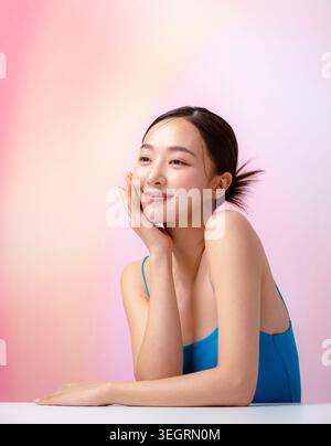 Eine junge asiatische Frau sitzt mit der Hand auf der Wange, lächelt und blickt direkt in die Kamera. Stockfoto