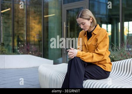 Junge Frau, die auf einer modernen städtischen Bank sitzt, mit nachdenklichem Ausdruck nach unten blickt, sich einsam und gestresst fühlt, während sie über Probleme im Freien nachdenkt Stockfoto