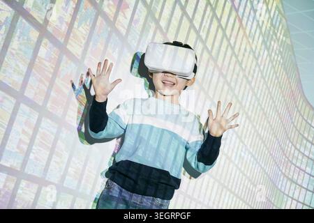 Ein Junge trägt ein Virtual-Reality-Headset und lächelt, umgeben von einer Stadtlandschaft an einer Wand. Stockfoto