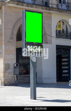 Totem-Kiosk mit grünem Display mit Chroma Key und LED-Digitaluhr, die 14:24 Uhr gegenüber der alten Gebäudefassade anzeigt. Urbane Umgebung Stockfoto