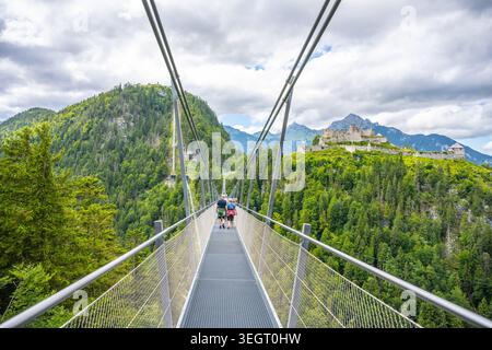 Besucher laufen auf der Highline 179, einer Hängebrücke in Österreich. Die Brücke verbindet eine alte Burgruine mit einer römischen Festung über eine breite Flussschlucht, die von Bergen und Bäumen umgeben ist. Stockfoto