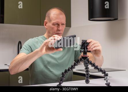 Senior man filmt mit Smartphone auf einem flexiblen Stativ am Tisch. Ausgereifter Mann nimmt Videos mit einem mobilen Gerät auf einem verstellbaren Ständer in Innenräumen auf. Startseite Stockfoto