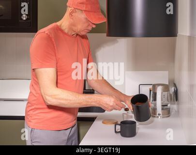 Reifer Mann gießt heißes Wasser aus dem Wasserkocher in eine Tasse an der Küchentheke. Erwachsene männlich bereitet Getränke mit Elektrogeräten im Innenbereich zu. Alltags-Haushalt l Stockfoto