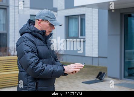 Ein reifer Mann benutzt ein Smartphone, während er in der Nähe des Hofes eines Wohngebäudes steht. Senior männlich überprüft mobiles Gerät im Freien neben der Bank des Wohnkomplexes Stockfoto
