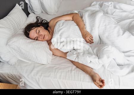 Weiße Frau mittleren Alters mit langen dunklen Haaren schläft friedlich in einem gemütlichen Bett, umgeben von weißen Kissen und Decken. Stockfoto