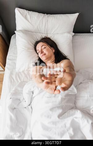 Frau mit langen dunklen Haaren dehnt sich im Bett. Sie ist mit einer weißen Decke bedeckt und wirkt entspannt in einem modernen Schlafzimmer. Stockfoto