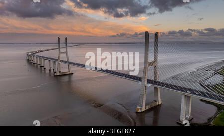 Aus der Vogelperspektive auf die Vasco da Gama-Brücke, die den Tejo überspannt, unter einem dramatischen Sonnenuntergangshimmel mit bunten Wolken in Lisboa, Portugal. Stockfoto
