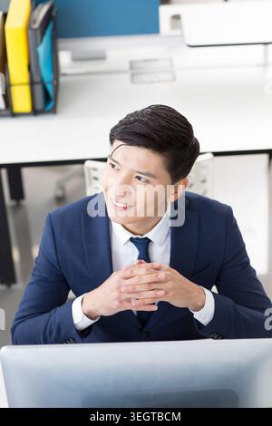 Ein junger asiatischer Geschäftsmann in einem marineblauen Anzug sitzt an einem Schreibtisch in einem modernen Büro und blickt auf einen Computerbildschirm mit einem fokussierten Ausdruck. Stockfoto