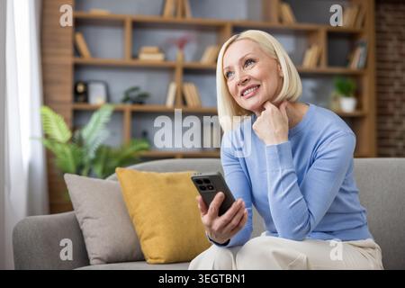 Blonde, reife Frau, die auf einem bequemen Sofa sitzt, ein Smartphone hält und nachdenklich wegblickt. Sie genießt einen Moment der Entspannung und Konnektivität in ihrem modernen Wohnzimmer Stockfoto