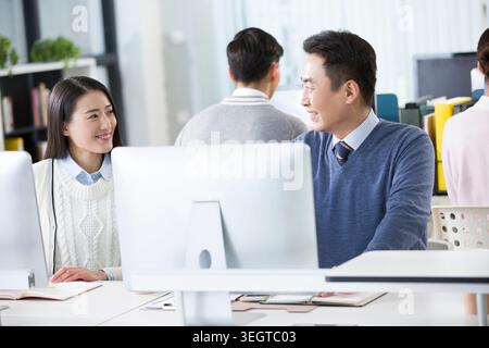 Eine junge asiatische Frau und ein asiatischer Mann mittleren Alters lächeln einander an, während sie an einem modernen Schreibtisch arbeiten. Stockfoto