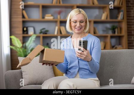 Seniorin sitzt auf einem Sofa, lächelt und hält einen Lieferkarton, während sie Artikel auf ihrem Smartphone überprüft, und genießt bequemen Online-Einkauf und Lieferservice zu Hause Stockfoto