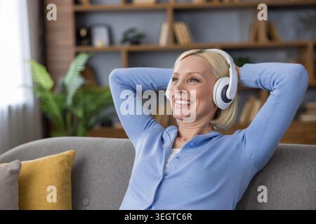Die reife blonde Frau lächelt und entspannt sich zu Hause auf einem Sofa, genießt ihre Freizeit, hört Musik oder ein Hörbuch über weiße Kopfhörer mit Händen hinter dem Kopf Stockfoto