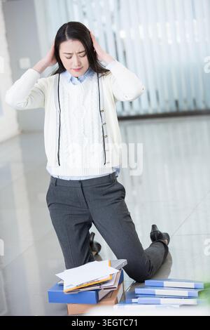 Eine gestresste Büroangestellte kniet auf einem unordentlichen Schreibtisch und hält ihren Kopf in den Händen. Stockfoto