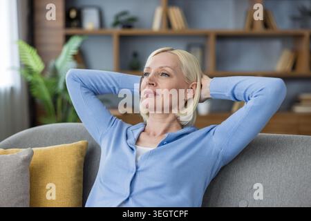 Reife blonde Frau, die bequem auf einem modernen Sofa sitzt und mit den Händen hinter dem Kopf einen Moment der Entspannung und Ruhe in einem gemütlichen Zuhause genießt Stockfoto