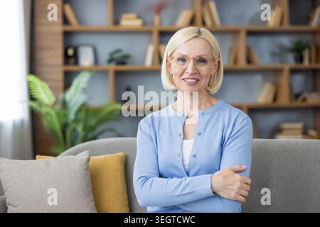 Selbstbewusste, reife Frau mit bob-Haarschnitt und Brille, die mit überkreuzten Armen lächelt, sitzt auf einer Couch in einer gemütlichen Wohnumgebung mit Bücherregalen und Pflanzen Stockfoto