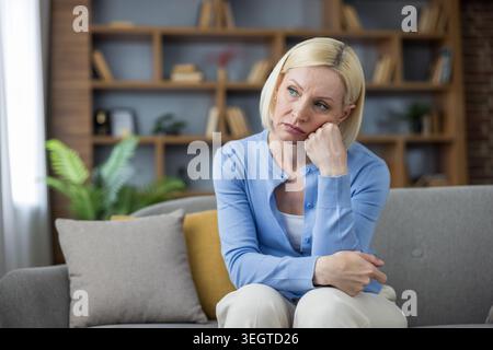 Blonde reife Frau sitzt auf einem Sofa in ihrem Wohnzimmer, fühlt sich traurig und einsam, denkt an Probleme, erlebt psychische Probleme oder Depressionen Stockfoto