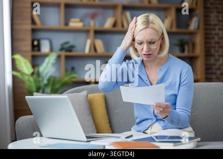 Gestresste Frau mittleren Alters, die Dokumente und Rechnungen überprüft, hat finanzielle Sorgen und Kopfschmerzen, während sie Haushaltsbudget und Schulden von ihrem Schreibtisch aus verwaltet Stockfoto
