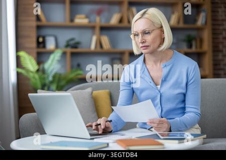 Selbstbewusste blonde Geschäftsfrau mit Brille und blauer Strickjacke, die auf einem Sofa sitzt, sich konzentriert, während sie an einem Laptop arbeitet und Dokumente in ihrem Heimbüro hält Stockfoto