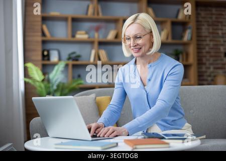 Blonde Geschäftsfrau mit Brille, die online arbeitet, auf dem Sofa sitzt, einen Computer für Fernarbeit oder Lernen benutzt, flexible Terminpläne und modernen digitalen Lebensstil genießt Stockfoto