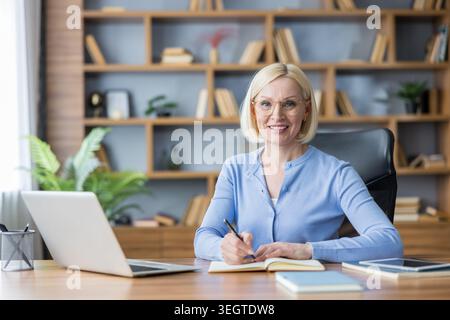 Blonde Geschäftsfrau mit Brille, die an einem Holzschreibtisch in einer Büroumgebung sitzt und selbstbewusst lächelt, während sie in einem Notebook schreibt, mit Laptop und Tablet in der Nähe Stockfoto